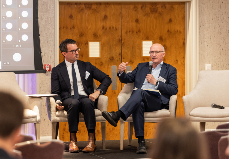 Two panel speakers seated on stage, holding microphones and speaking during a conference session, with chairs, a screen, and wooden double doors visible behind them.