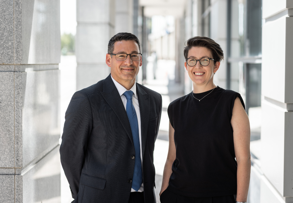 Two people in business attire stand side by side in a bright, modern outdoor corridor with stone columns and glass walls, both facing the camera and wearing glasses; one is in a dark suit and tie, the other in a sleeveless black top.